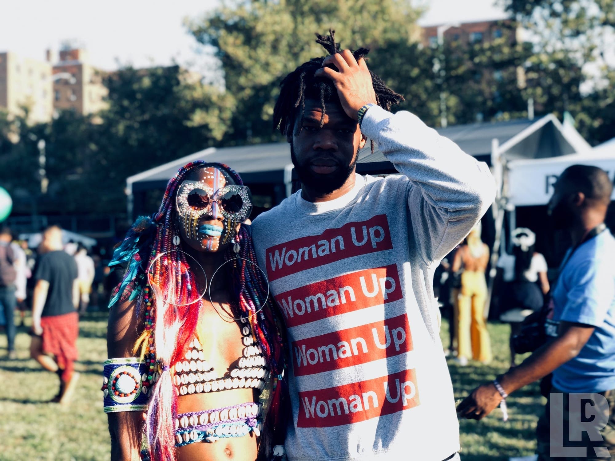Two festival-goers posing together, one in vibrant costume with shells and feathers, the other in a Woman Up sweatshirt