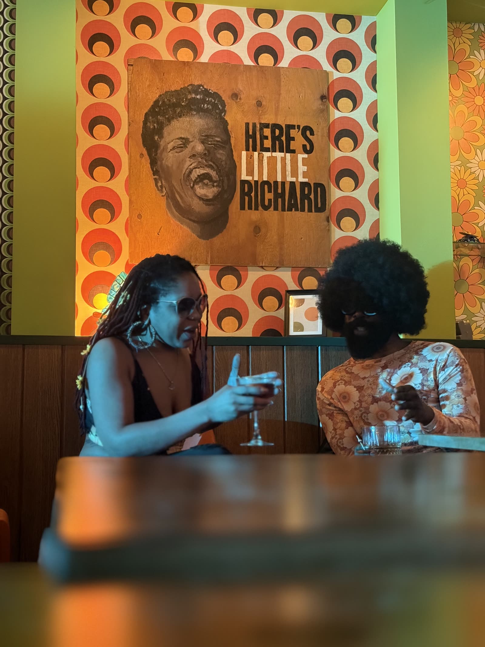 Two people talking in a booth with drinks, beneath a painted portrait of Little Richard on plywood.