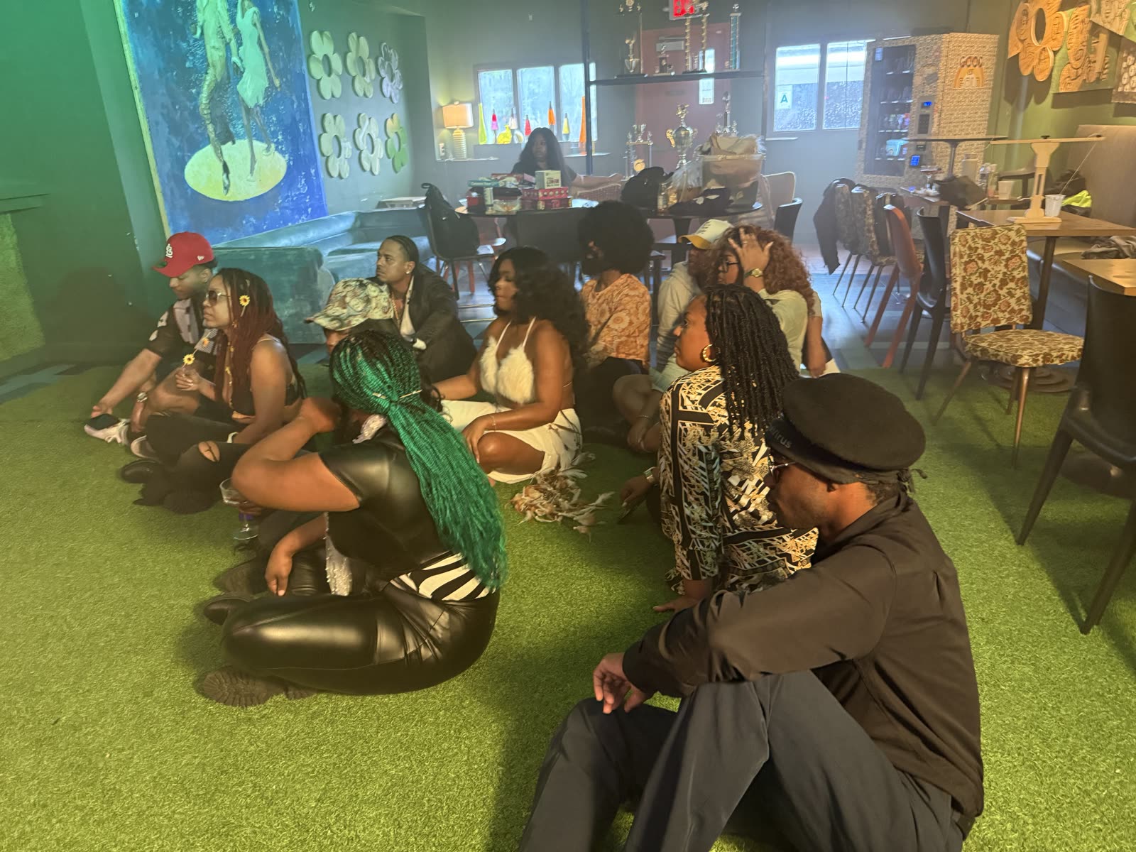 Cast members sitting together on a green floor inside the bar between takes.