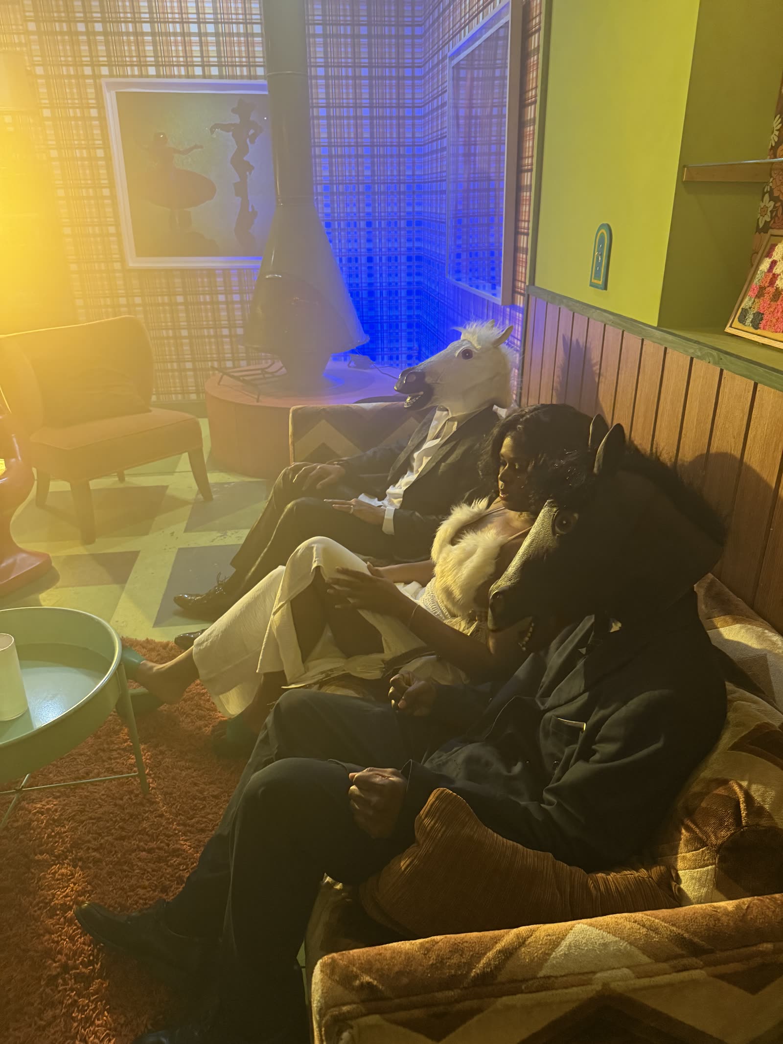 Two performers in black and white horse masks sitting on a couch with other cast members.
