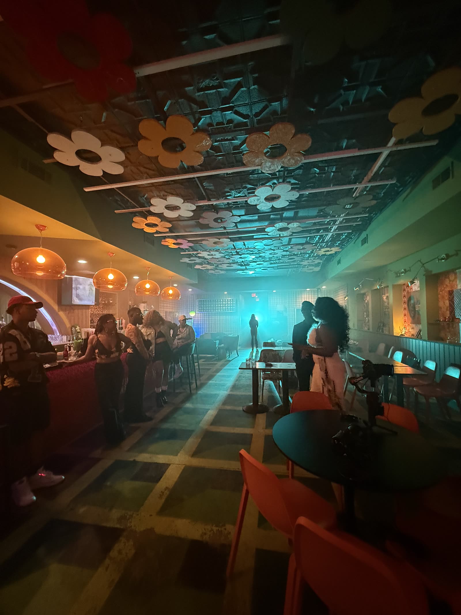 Wide interior of the bar under teal and orange light, with large fabric flowers fixed to the ceiling and a haze across the room.